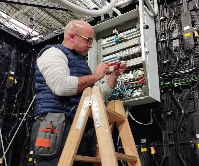 Instalador conectando un cuadro detrás de una videopantalla, en un pabellón