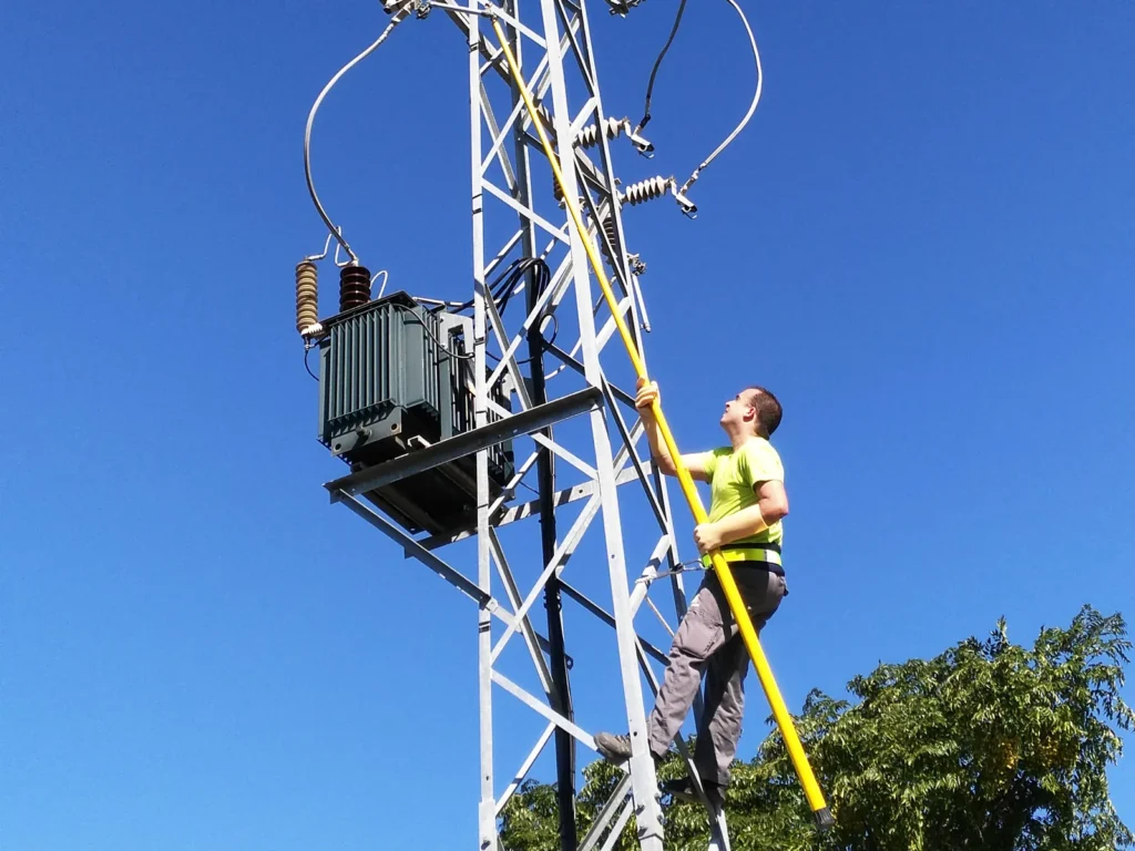 Técnico trabajando con una pértiga junto a un transformador montado en un apoyo de línea aérea