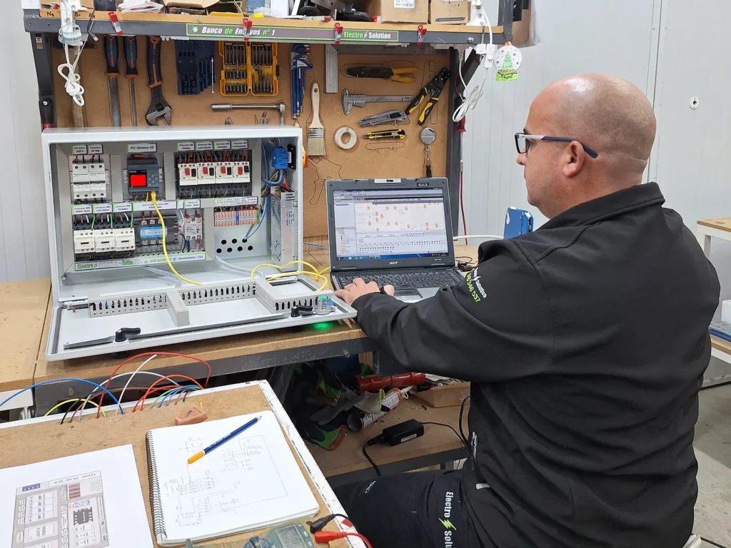 Técnico de ElectroSolution en el banco de ensayos, con un cuadro abierto conectado al portátil y los esquemas sobre la mesa
